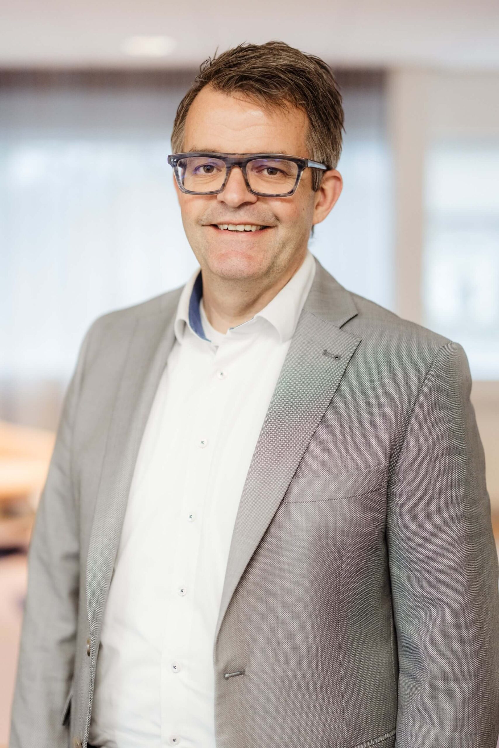 Man met bril in grijs colbert voor een onscherpe kantoorachtergrond.