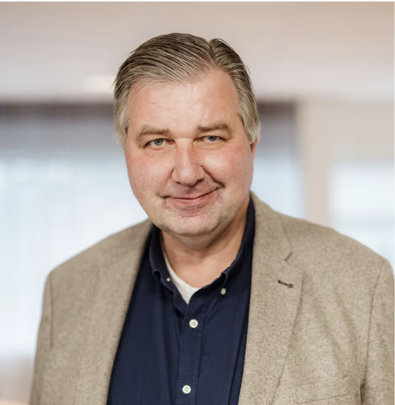 Man in beige colbert voor een onscherpe kantoorachtergrond.