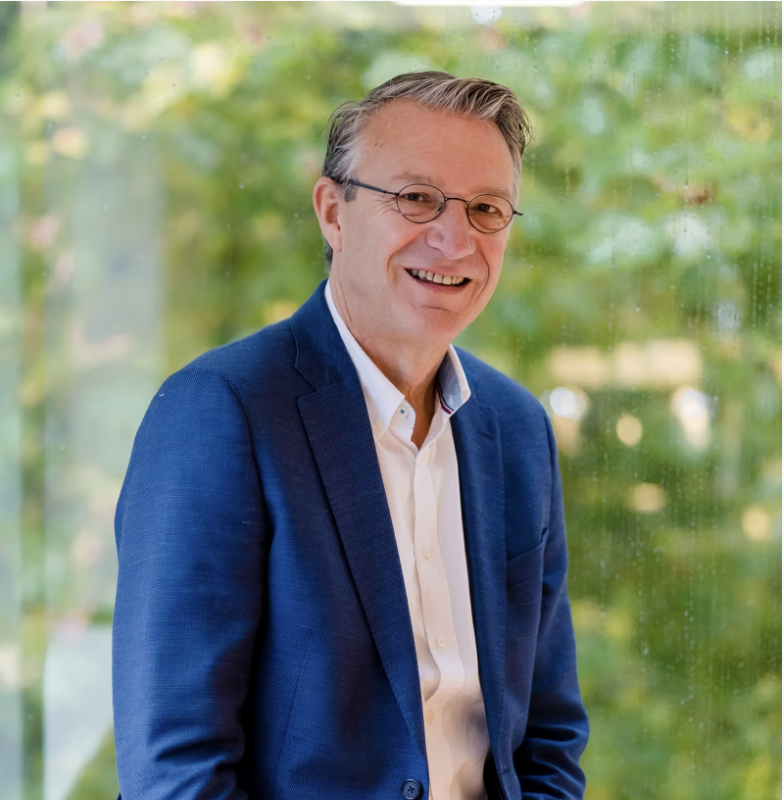 Lachende man met bril en blauw colbert voor een groene, onscherpe achtergrond.