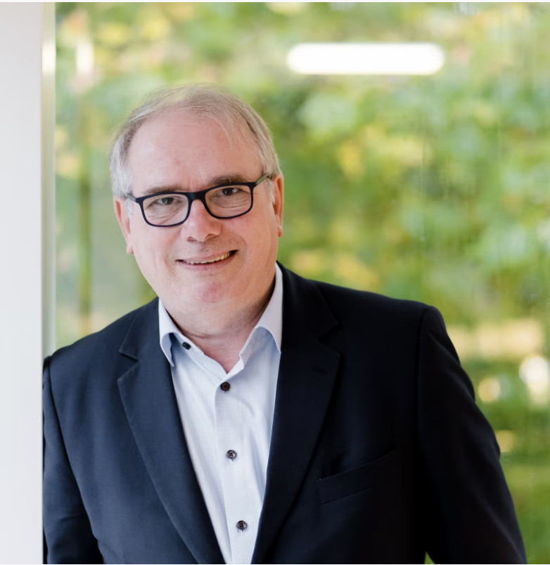 Lachende man met bril en zwart colbert voor een groene, onscherpe achtergrond.