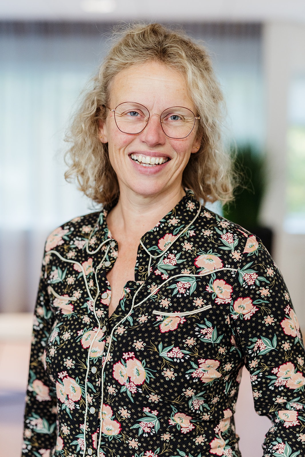 Vrouw met bril en bloemetjesblouse voor een onscherpe kantoorachtergrond.