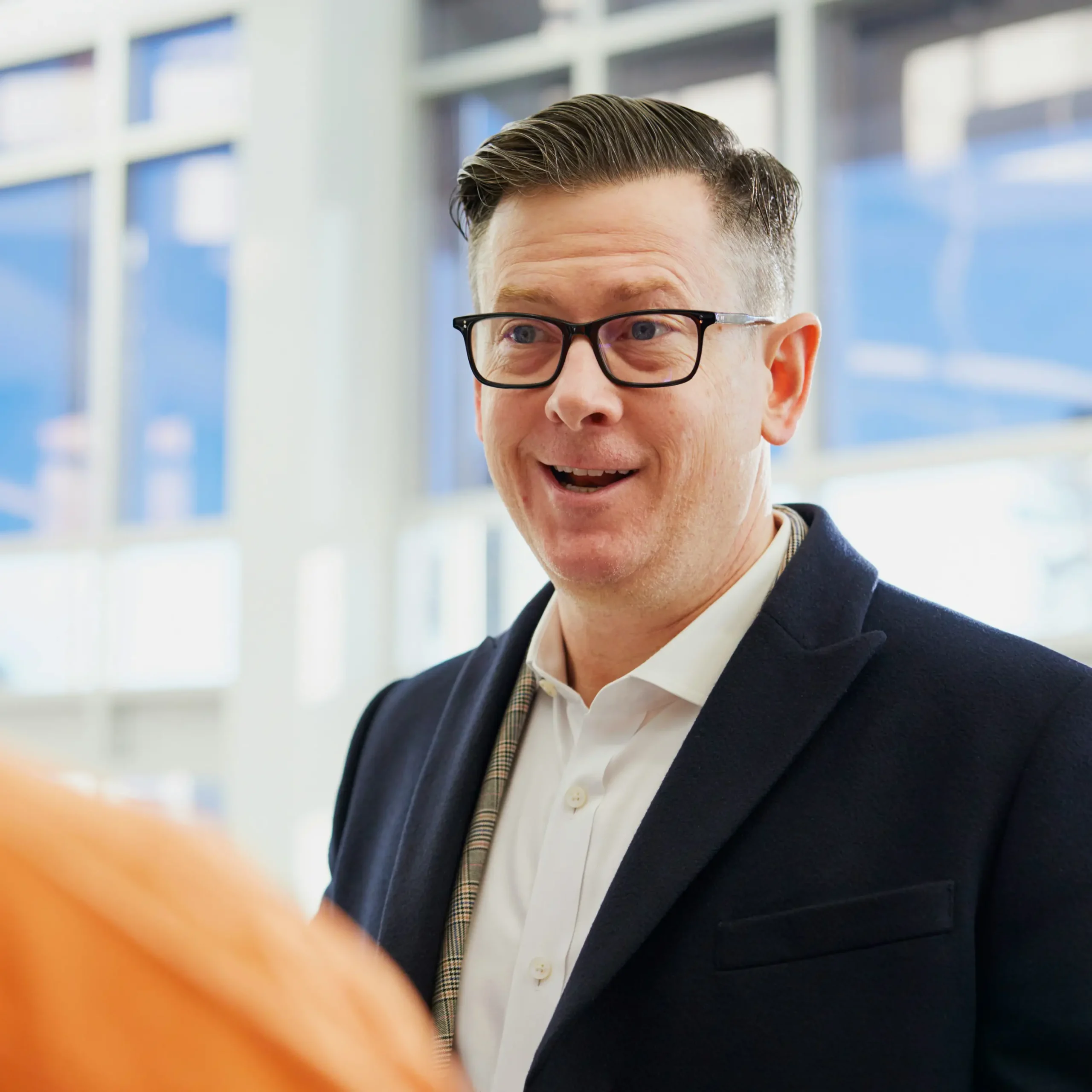 Lachende man met bril in een donker colbert in gesprek op een lichte kantoorlocatie.