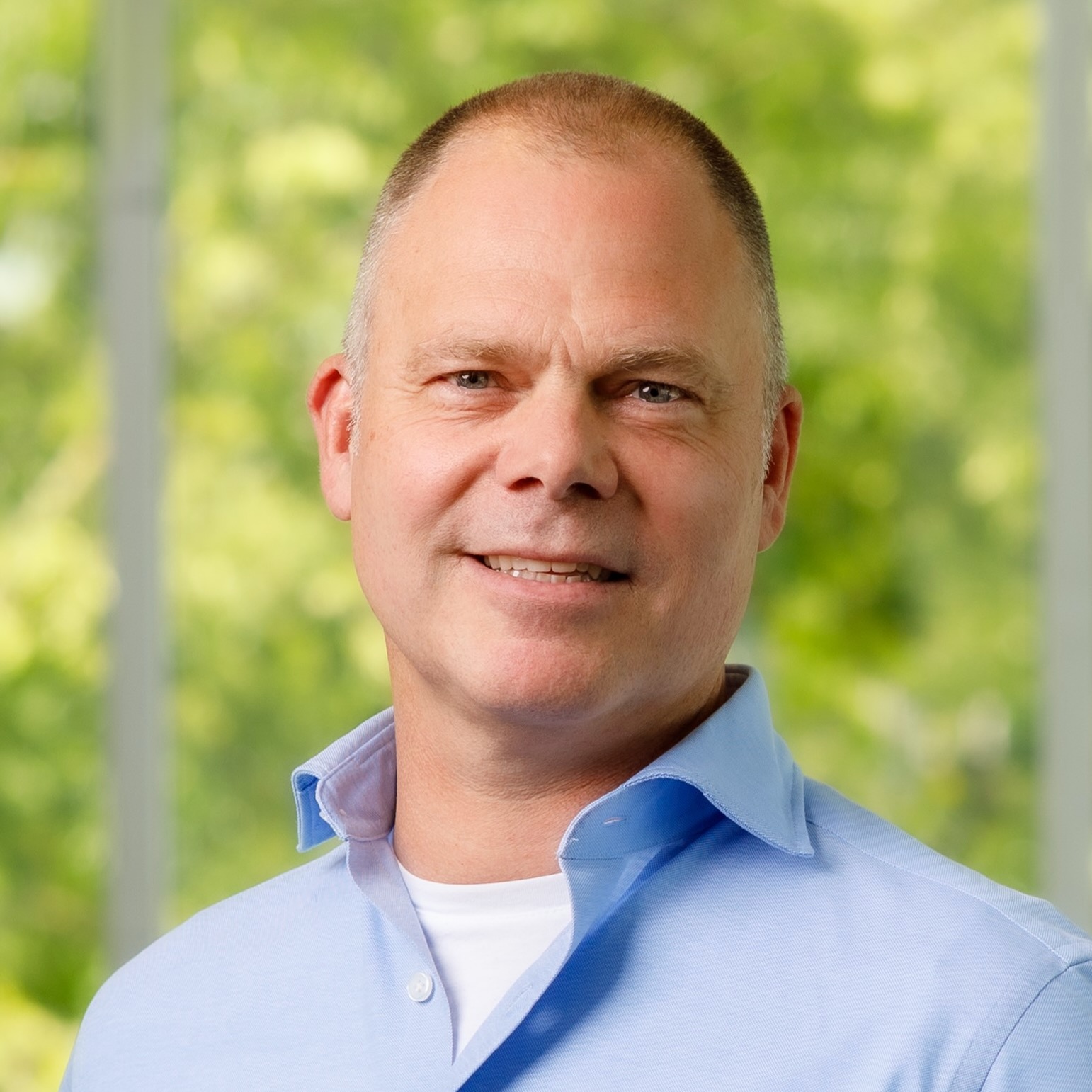 Lachende man in een lichtblauw overhemd voor een groene, onscherpe achtergrond.