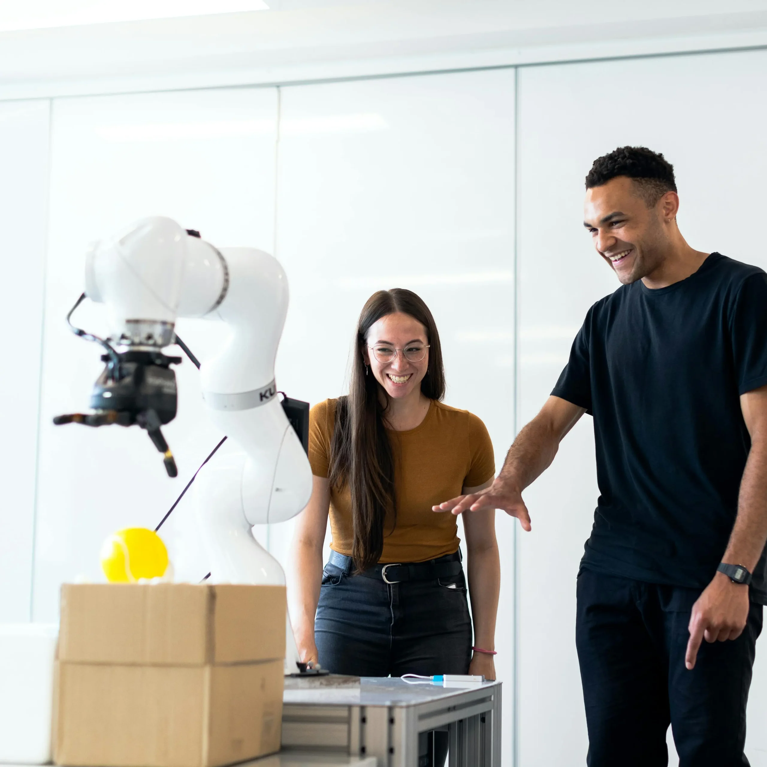 Twee mensen staan lachend naast een witte robotarm in een werkruimte.