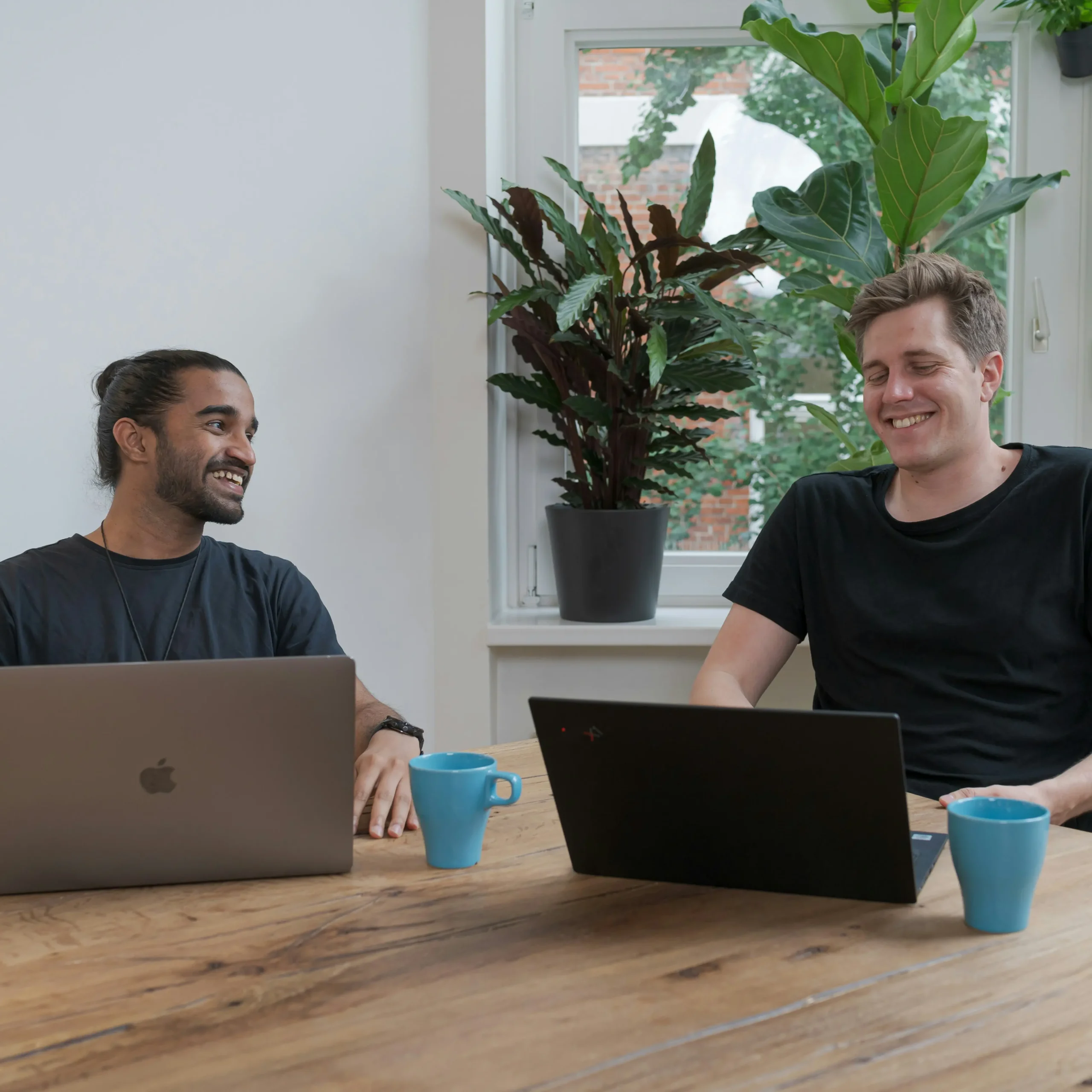 Twee mannen met laptops praten en lachen aan een houten tafel met koffiekopjes.