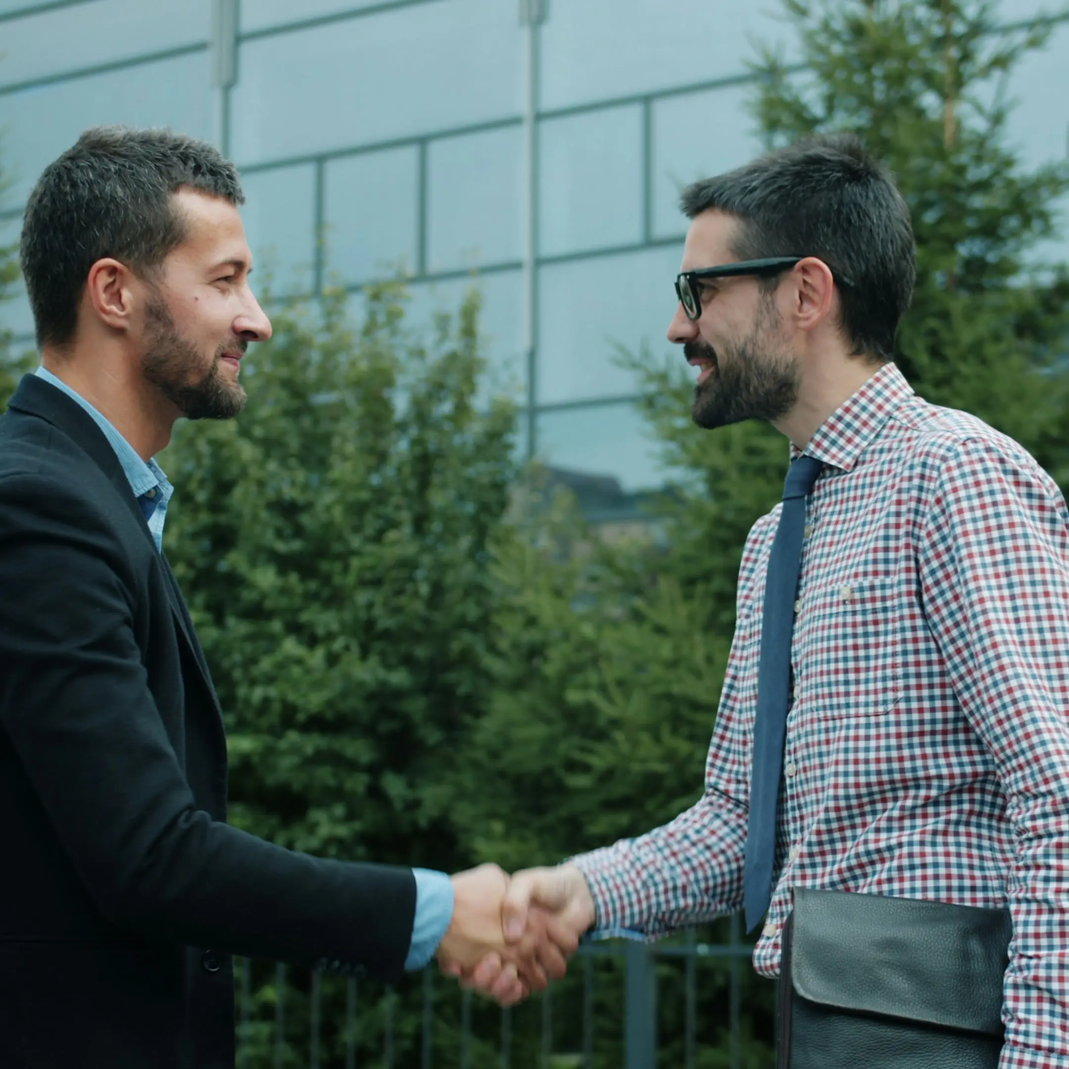 Twee mannen in zakelijke kleding schudden elkaar de hand buiten voor een modern gebouw.