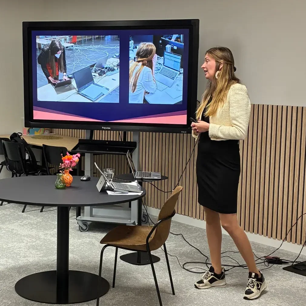 Vrouw geeft een presentatie bij een groot scherm terwijl een man op de voorgrond een foto maakt met zijn telefoon.