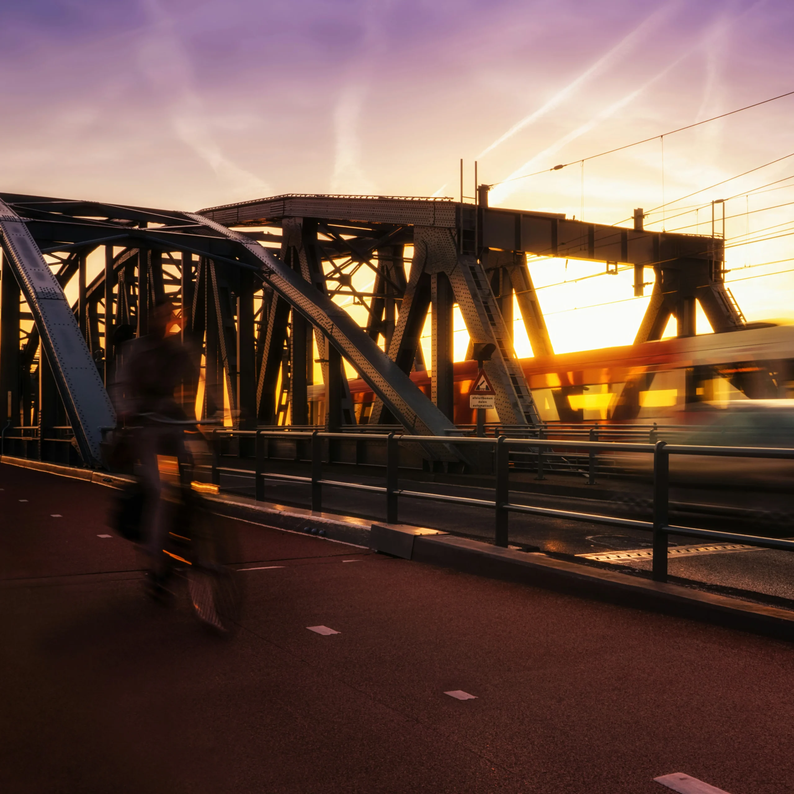 Fietser en passerende trein op een stalen spoorbrug in warm avondlicht.